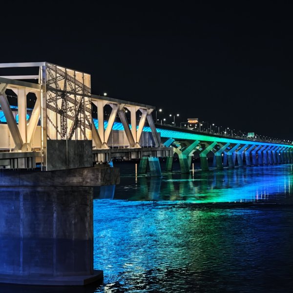 Passerelle pi&eacute;tonne moderne sur l&rsquo;&Icirc;le-des-S&oelig;urs, dot&eacute;e de structures m&eacute;talliques blanches en arche et de panneaux textur&eacute;s perfor&eacute;s, avec un rev&ecirc;tement en bois et plusieurs personnes marchant ou discutant dans un am&eacute;nagement paysager urbain.