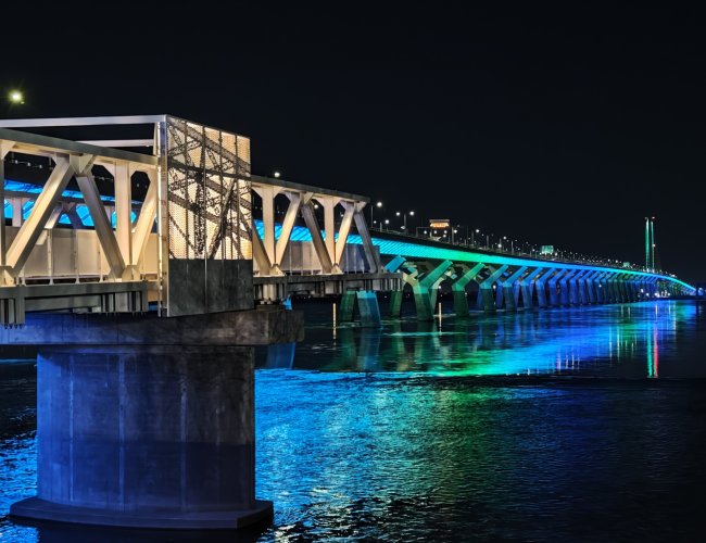 Passerelle pi&eacute;tonne moderne sur l&rsquo;&Icirc;le-des-S&oelig;urs, dot&eacute;e de structures m&eacute;talliques blanches en arche et de panneaux textur&eacute;s perfor&eacute;s, avec un rev&ecirc;tement en bois et plusieurs personnes marchant ou discutant dans un am&eacute;nagement paysager urbain.